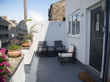 An outdoor patio with three chairs a small table flower pots on a ledge black door and white walls at Seashell Cottage in Amble-By-The-Sea