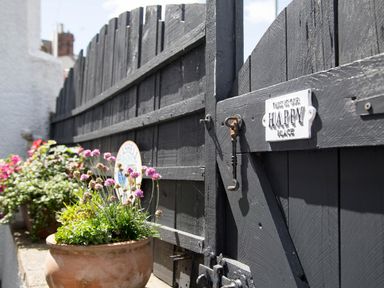 A black wooden gate with a metal sign and a potted plant with flowers at Seashell Cottage in Amble-By-The-Sea