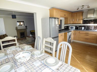 A kitchen and dining area with wooden cabinets a dining table set with plates and glasses and a living room with a fireplace in the background at Oakdene in Neyland