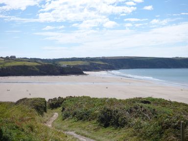 A coastal landscape with a sandy beach people walking on the beach and grassy cliffs in the background