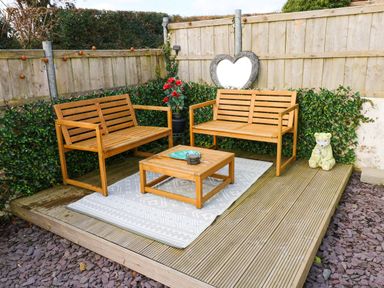 A wooden deck with two wooden benches a wooden coffee table a patterned rug potted artificial red flowers a heart-shaped decoration and a bear statue at Oakdene in Neyland