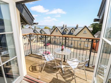 A balcony with metal chairs and a table overlooking yellow houses and a distant sea view at 24 Rhodewood House in Saundersfoot