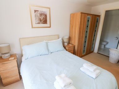 A bedroom with a double bed with blue bedding and towels on it two wooden nightstands with lamps a wooden wardrobe and a bathroom visible through an open door at 24 Rhodewood House in Saundersfoot