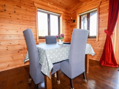 A dining area with a table covered by a star-patterned tablecloth and four chairs with slipcovers in a wooden room with windows and red curtains at Willow Lodge in Lanivet near Bodmin