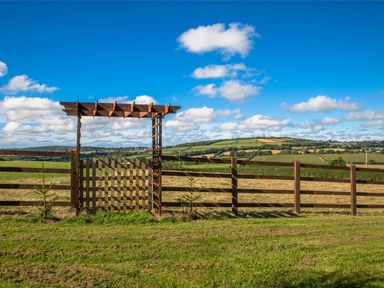 A wooden gate with a pergola and fence overlooking green fields and hills under a blue sky at Holly Lodge in Lanivet near Bodmin