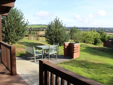 An outdoor patio area with a wooden table and chairs near a brick barbecue and a garden gate surrounded by grass and trees at Holly Lodge in Lanivet near Bodmin