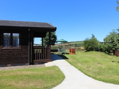 A wooden cabin with a patio table and umbrella on a lawn with a paved path and trees at Holly Lodge in Lanivet near Bodmin
