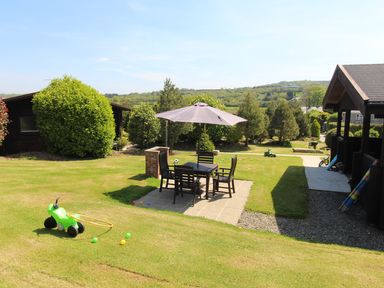 An outdoor garden area with a patio table and chairs under an umbrella and children's toys scattered on the grass at Hazel Lodge Lanivet near Bodmin