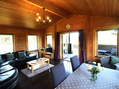 A wooden living room with black leather sofas a wooden coffee table a dining table with chairs and flowers at Hazel Lodge Lanivet near Bodmin