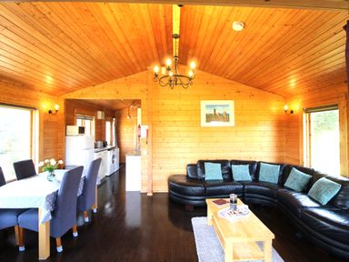 A wooden interior living room with a dining table and chairs next to a kitchen and a black sectional sofa with cushions at Hazel Lodge in Lanivet near Bodmin