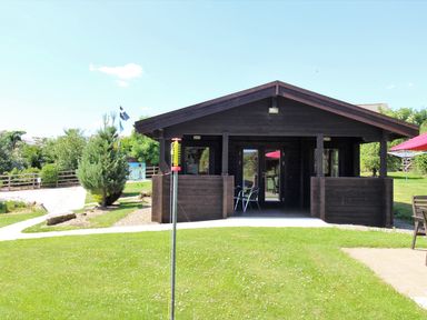 A wooden cabin with a porch and chairs on grass with trees and flags in the background at Hazel Lodge in Lanivet near Bodmin