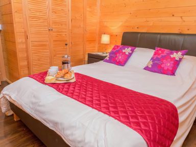 A bedroom with a bed with white sheets red runner purple floral pillows a side table with a lamp and a wooden wardrobe at Hazel Lodge in Lanivet near Bodmin