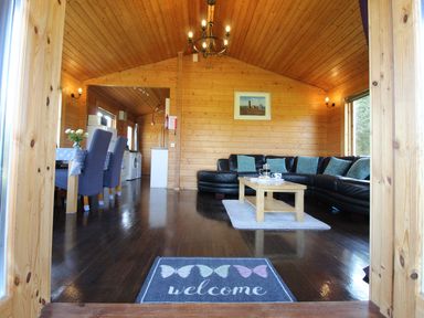 A wooden living room with a black sectional sofa and coffee table near a dining area with blue chairs at Hazel Lodge in Lanivet near Bodmin