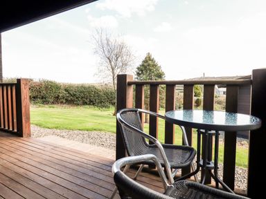 A wooden outdoor deck with a glass round table and two metal and wicker chairs overlooking a grassy yard with trees and hedges at Cherry Lodge Lanivet near Bodmin