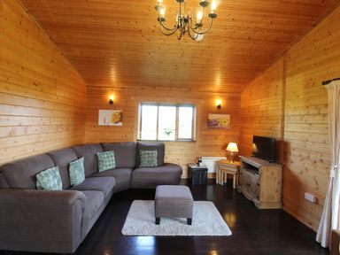 A living room with a gray corner sofa a small ottoman a tv on a wooden cabinet and wooden walls at Cherry Lodge in Lanivet near Bodmin