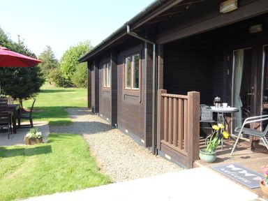 The exterior of a wooden cabin with a small porch furnished with a table, chairs, and potted flowers at Cherry Lodge in Lanivet near Bodmin