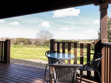 A covered wooden porch with a round glass table and four black chairs overlooking a grassy yard at Maple Lodge in Lanivet near Bodmin