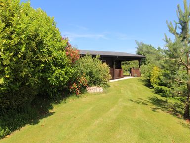 A grass lawn with bushes and trees surrounding a wooden cabin at Maple Lodge in Lanivet near Bodmin