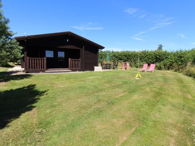 A wooden cabin with a veranda in a grassy yard with outdoor chairs a table an umbrella and two pink lounge chairs at Maple Lodge in Lanivet near Bodmin