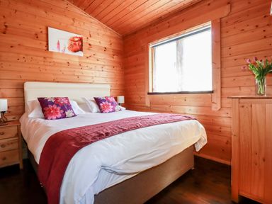 A bedroom with a double bed with white and red bedding wooden walls a window and a wooden dresser with flowers at Cedar Lodge Lanivet near Bodmin