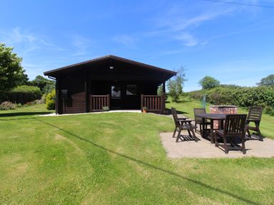 A wooden lodge with a small porch and outdoor dining table with chairs on a paved area on a grassy lawn at Cedar Lodge in Lanivet near Bodmin