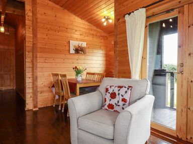 A wooden dining area with chairs and a table with flowers beside an armchair near a glass door at Cedar Lodge Lanivet near Bodmin