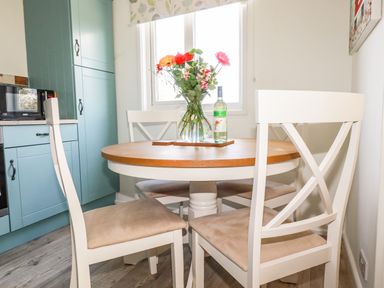 A kitchen dining area with a round table and four white chairs near a window with flowers in a vase and a bottle on the table at Hunrosa St Day