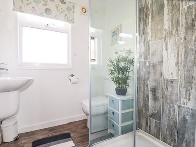 A bathroom with a white sink a window a toilet with a small cabinet and a plant next to a shower with patterned wall tiles at Hunrosa in St Day