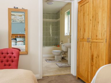 A bedroom with a wooden wardrobe red armchair and a mirror showing a bed and an adjacent bathroom with a toilet sink and bathtub at Richmond Cottage in Burythorpe near Malton