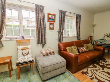 A living room with a brown leather sofa and cushions a grey ottoman with blankets and chairs near windows with curtains at Richmond Cottage in Burythorpe near Malton