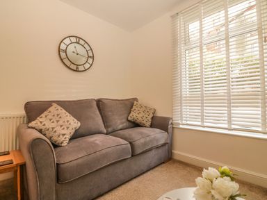 A room with a grey sofa with patterned cushions a wooden side table a wall clock white blinds on the window and flowers on a table at Sunvale in Teignmouth