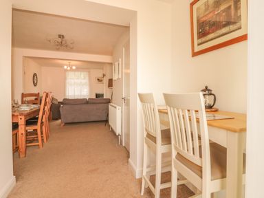 A hallway with a small wooden table and two white chairs leading to a dining area with wooden table and chairs and a living room with a grey sofa at Sunvale in Teignmouth