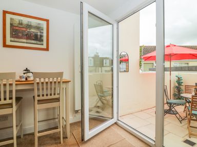 A dining area with two chairs and a table next to an open glass door leading to a patio with red umbrellas and outdoor chairs at Sunvale in Teignmouth
