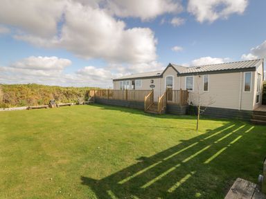 A mobile home with a wooden deck and steps on a lawn with benches and a small tree at Coed Llai Lodge in Trearddur Bay