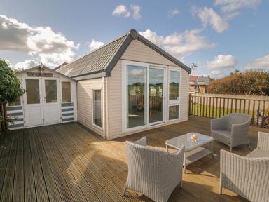 An outdoor wooden deck with patio chairs and a table alongside a small beige building and a white shed at Coed Llai Lodge in Trearddur Bay
