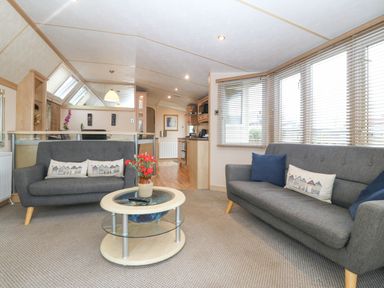 A living room with two gray sofas a round glass coffee table with flowers and a kitchen in the background at Coed Llai Lodge in Trearddur Bay