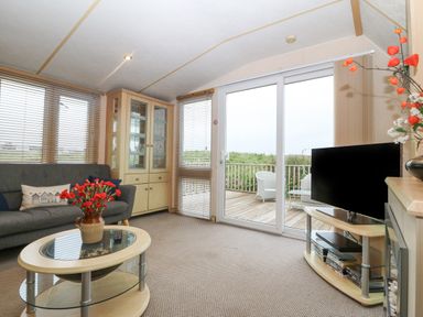 A living room with a sofa glass cabinet coffee table television and sliding doors leading to a wooden deck at Coed Llai Lodge in Trearddur Bay