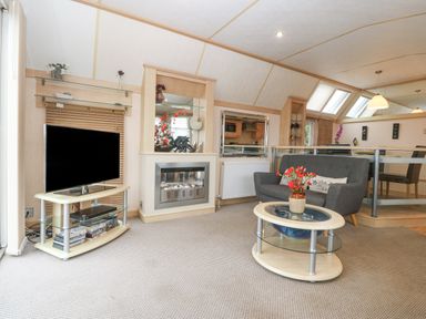 A living area with a gray sofa, a glass coffee table with a flower vase, a television on a stand, and a fireplace at Coed Llai Lodge in Trearddur Bay