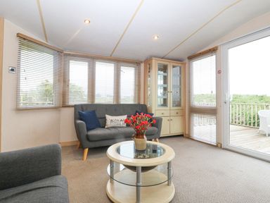 A living room with sofas a round coffee table with flowers and a glass cabinet by windows at Coed Llai Lodge in Trearddur Bay