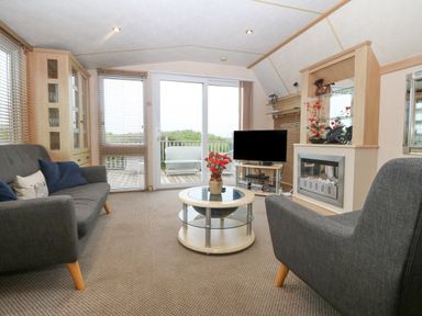 A living room with two gray armchairs a round glass coffee table a TV on a stand a fireplace a wooden cabinet and sliding glass doors at Coed Llai Lodge in Trearddur Bay