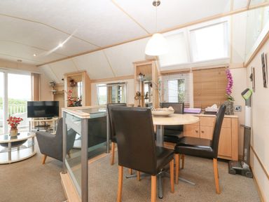A dining area with a round table and four chairs next to a living room with a TV and armchair at Coed Llai Lodge in Trearddur Bay
