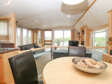 A dining area with a round table and black chairs looking out to a living area with a sofa and glass doors at Coed Llai Lodge in Trearddur Bay