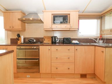 A kitchen with wooden cabinets a gas stove oven microwave kettle toaster and a stainless steel sink at Coed Llai Lodge in Trearddur Bay