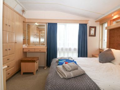 A bedroom with a bed with gray and white bedding towels on the bed a wooden wardrobe a dressing table with a mirror and a stool blue curtains and windows at Coed Llai Lodge in Trearddur Bay