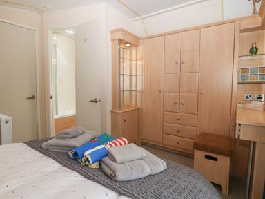 A bedroom with a bed and folded towels on it wooden wardrobes a small wooden stool and a desk with shelves at Coed Llai Lodge in Trearddur Bay