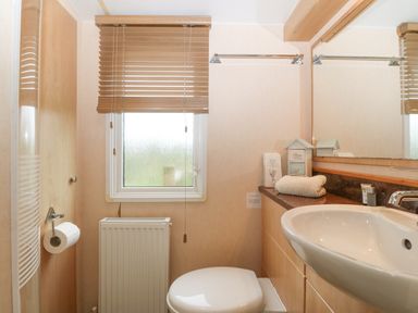 A bathroom with a sink toilet window with blinds and countertop with towel and small decorative items at Coed Llai Lodge in Trearddur Bay