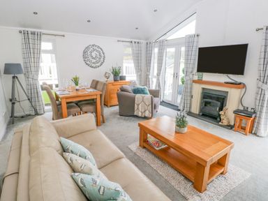 A living room with beige sofa, wooden coffee table with plant, dining table with chairs, armchair, wall-mounted TV above fireplace and patterned curtains at The Lookout in Stepaside