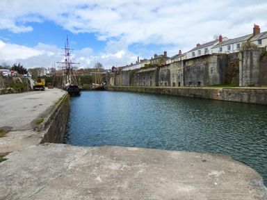 A dock with moored tall ships beside a stone wall and houses in Charlestown