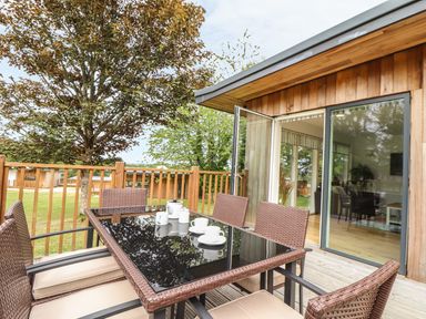 An outdoor wooden deck with a glass table and six wicker chairs next to a building with large glass doors at Birch in Truro