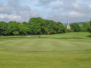 A golf course green with a flag and trees in the background near a church with a spire at Birch in Truro